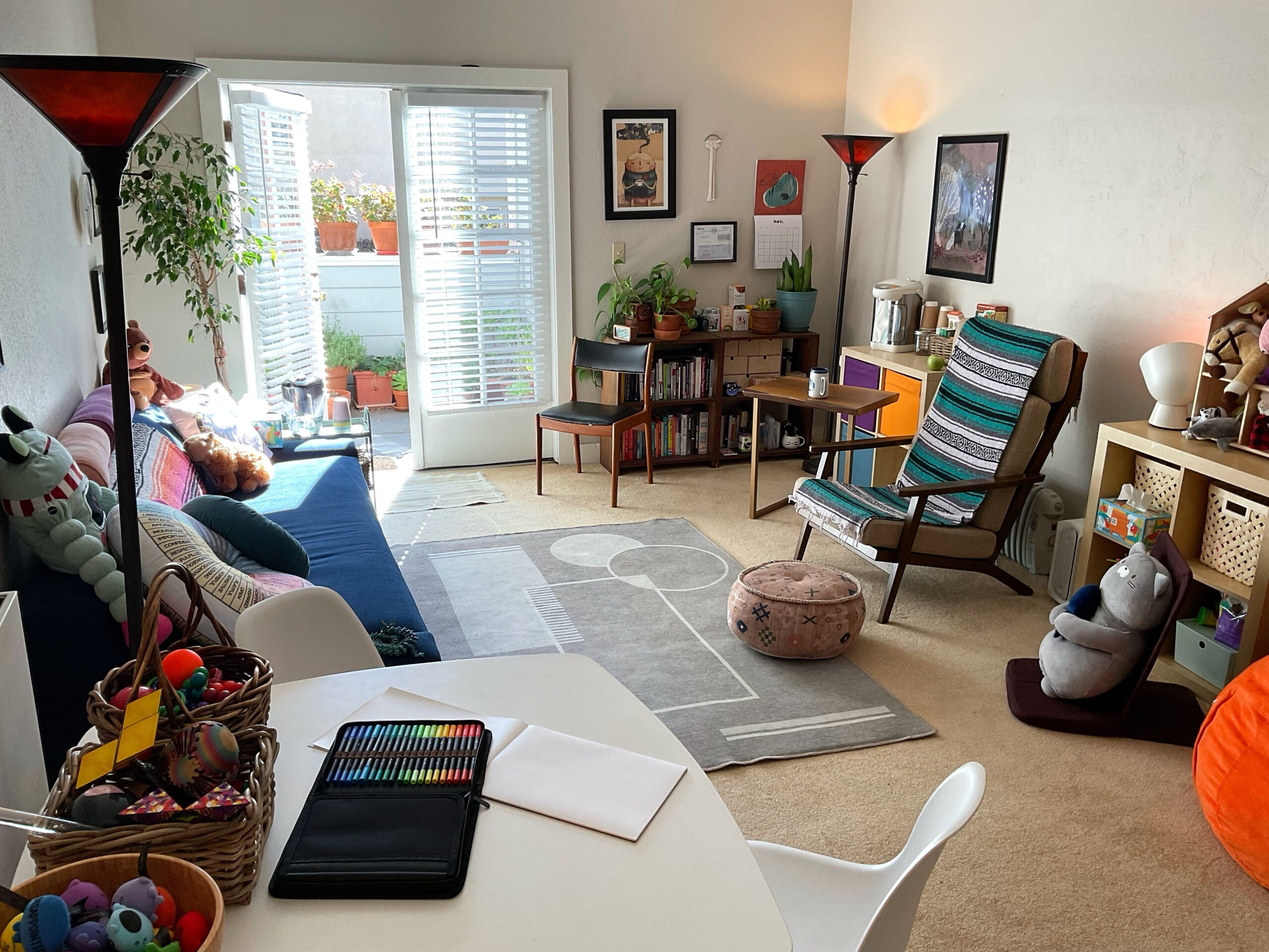 A photo of a large, bright, sunny therapy office with a large couch, a table with art supplies, and a bean bag.